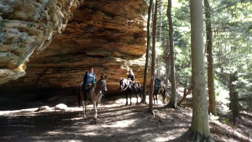 Two riders on horseback explore a forested area beneath a rocky overhang. Sunlight filters through the trees.