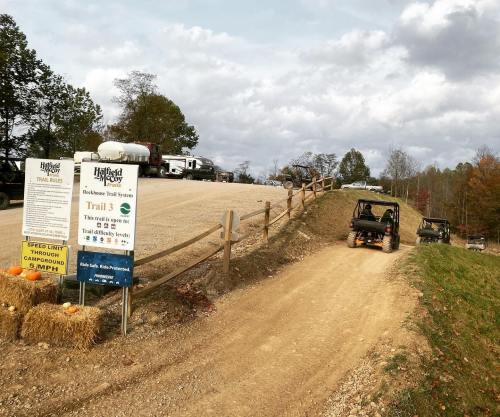 A dirt trail leading into a wooded area, with a sign for Hatfield McCoy Trails and parked off-road vehicles nearby.