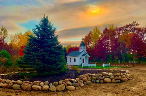 A small white chapel surrounded by colorful autumn trees and a stone border under a vibrant sky.