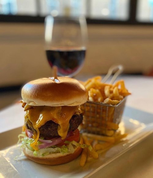 A juicy cheeseburger with lettuce, tomato, and onion, served with fries and a glass of red wine in the background.
