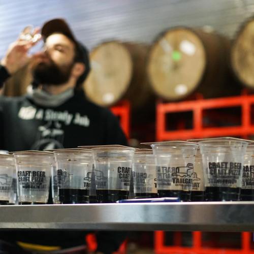 A person drinks from a cup in a brewery, with several cups of dark liquid lined up on a table in front.