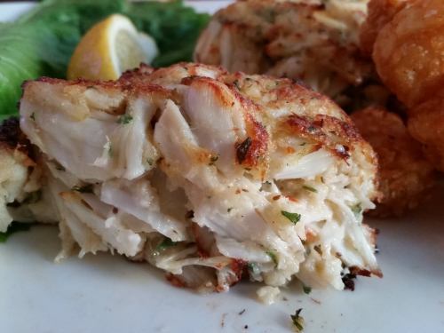 Close-up of a golden-brown crab cake, garnished with herbs and served with a lemon wedge and lettuce.