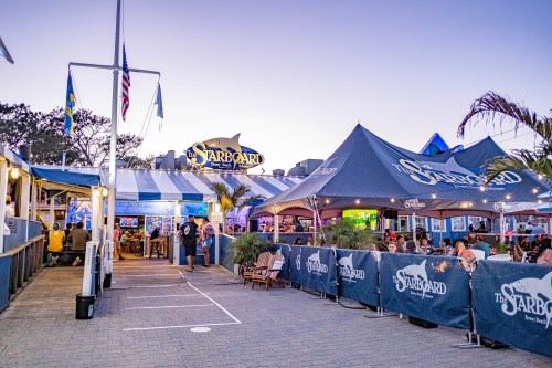 Outdoor dining area of a restaurant named "Starboard," with flags and a lively atmosphere at dusk.