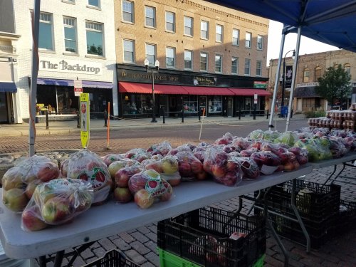A table at a market displays bags of apples, with shops and buildings in the background.