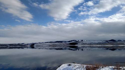 A serene landscape featuring a calm lake reflecting snowy hills under a cloudy sky.