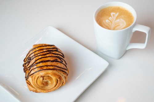 A chocolate-drizzled pastry on a white plate next to a cup of latte with a decorative foam design.
