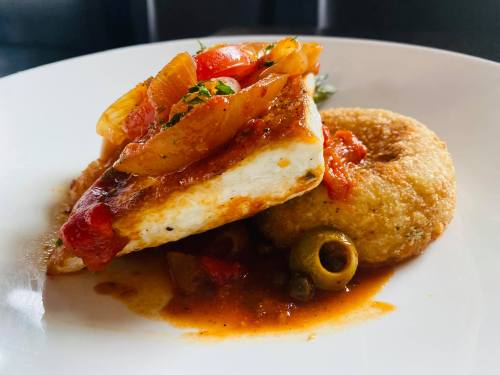 A plate featuring a piece of grilled fish topped with sautéed onions and peppers, served with a round fried dough.