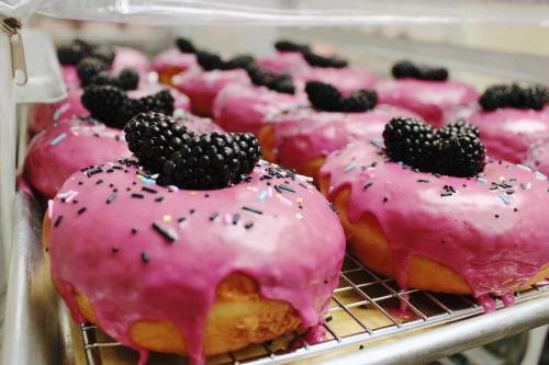 A tray of donuts with vibrant pink icing, topped with blackberries and colorful sprinkles, cooling on a wire rack.