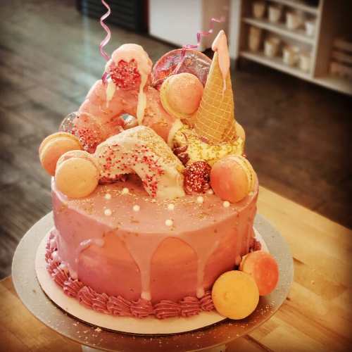 A colorful cake topped with macarons, an ice cream cone, and sprinkles, featuring a pink frosting and decorative elements.