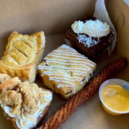 A box containing four desserts: a pastry, a brownie with frosting, a scone with icing, and a churro with dipping sauce.