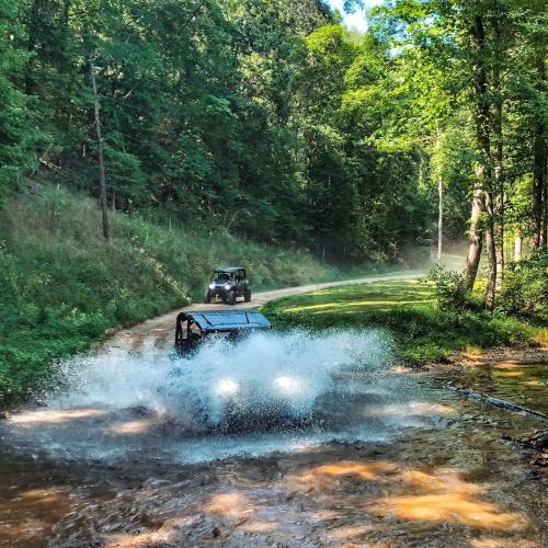 Two off-road vehicles splash through a muddy water crossing on a forested dirt path. Sunlight filters through the trees.