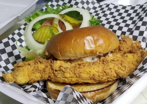 A crispy fried chicken sandwich on a bun, served with lettuce, tomato, pickles, and onion on a checkered paper.