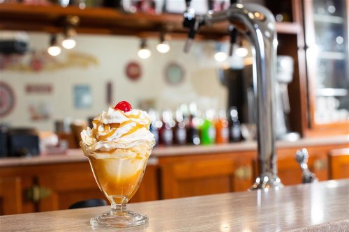 A glass dessert cup filled with ice cream, topped with whipped cream, caramel drizzle, and a cherry, on a bar counter.