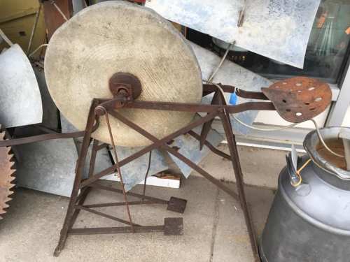 An old metal grinding wheel mounted on a rusted frame, with a paddle-like handle, beside various metal objects.