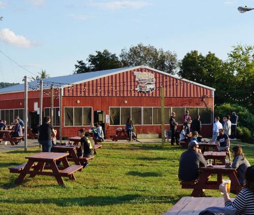 A red barn-style brewery with outdoor seating, people socializing, and a clear sky in the background.