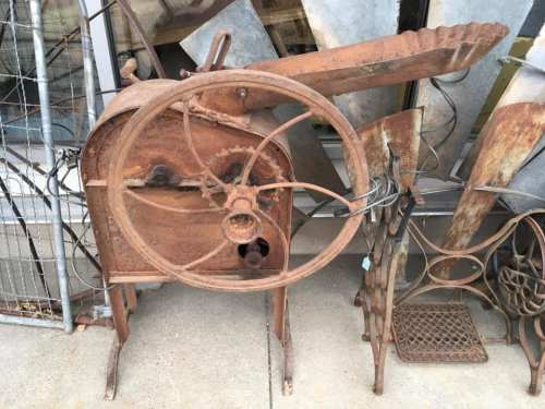 Rusty vintage machinery with a large wheel and metal components, displayed outdoors among other metal items.