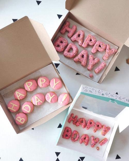 Three boxes of colorful donuts spelling "HAPPY BDAY!" with pink icing and sprinkles, set against a patterned background.