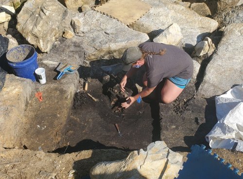 A person kneels in a rocky excavation site, carefully uncovering artifacts from the ground.