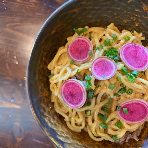 A bowl of creamy pasta topped with pink radish slices and green onions, set on a wooden table.