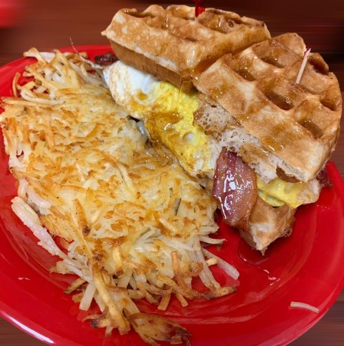 A red plate with a waffle sandwich filled with egg and bacon, alongside crispy golden hash browns.