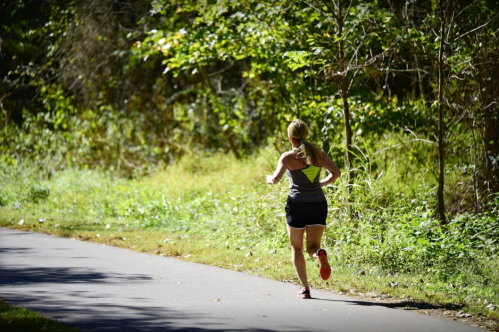 A woman runs along a tree-lined path on a sunny day, wearing a tank top and shorts.