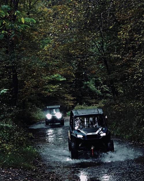 Two off-road vehicles drive through a forested trail, splashing through water under a canopy of autumn leaves.