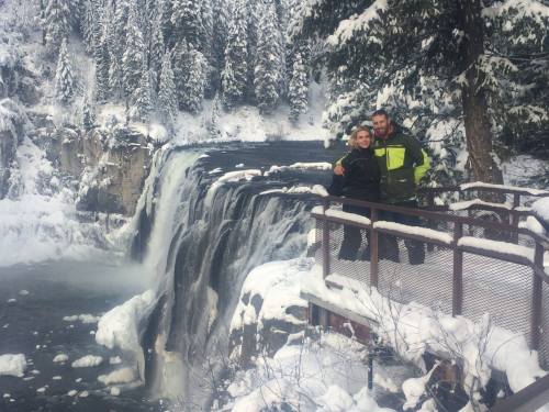 A couple stands on a snowy overlook by a waterfall, surrounded by snow-covered trees and a winter landscape.