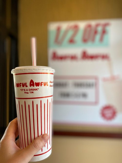 A hand holds a large drink cup with a straw, featuring a sign in the background advertising "1/2 OFF."