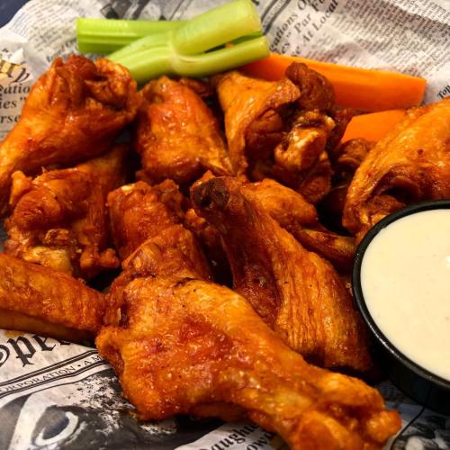 A platter of crispy chicken wings with celery, carrots, and a side of ranch dressing on a bed of newspaper.