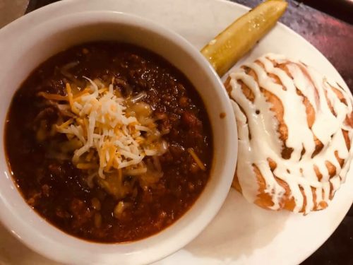 A bowl of chili topped with cheese, served with a cinnamon roll and a pickle on the side.