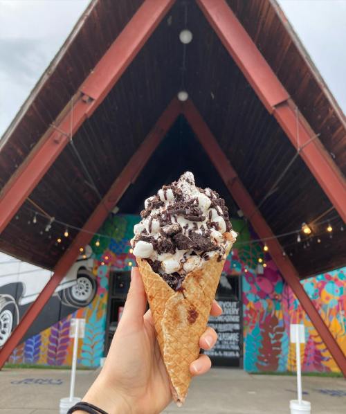 A hand holds a large ice cream cone topped with chocolate and marshmallows in front of a colorful mural and A-frame building.