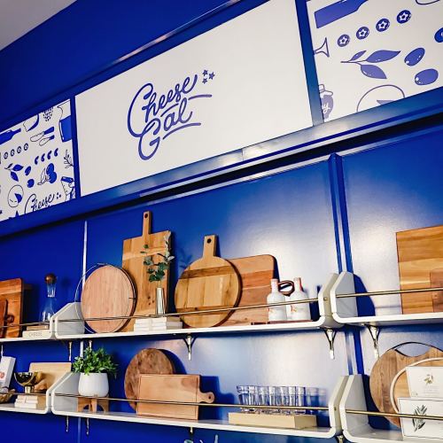 A blue wall display featuring wooden cutting boards, glassware, and a sign that reads "Cheese Gal."