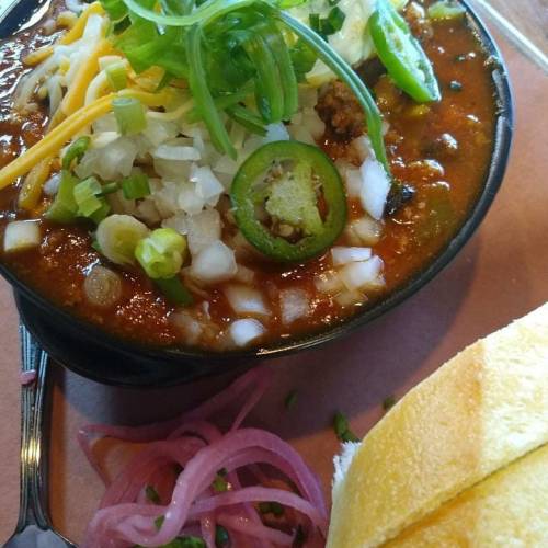 A bowl of chili topped with cheese, onions, jalapeños, and green onions, served with a side of bread and pickled onions.