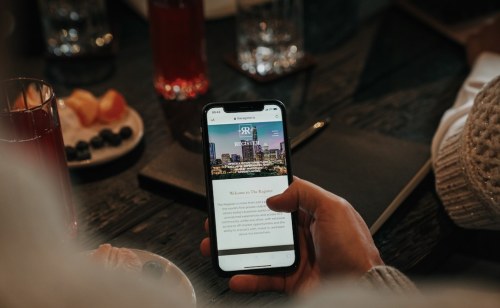 A person holding a smartphone displaying a website, with drinks and fruit on a table in the background.