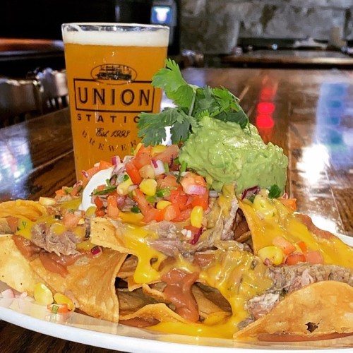 A plate of loaded nachos topped with guacamole, salsa, and cheese, alongside a glass of beer on a wooden table.