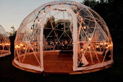 A cozy transparent dome with string lights, featuring wooden seating inside, set in a serene outdoor environment.
