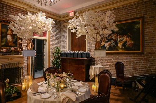 Elegant dining setup with floral centerpieces, soft lighting, and a brick wall backdrop in a cozy restaurant setting.