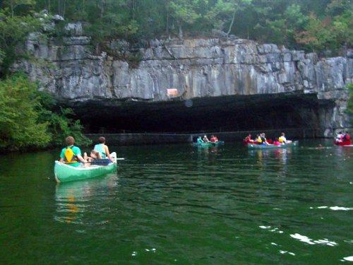Two canoes on a calm lake near a rocky cave entrance, surrounded by lush greenery and other boats in the distance.