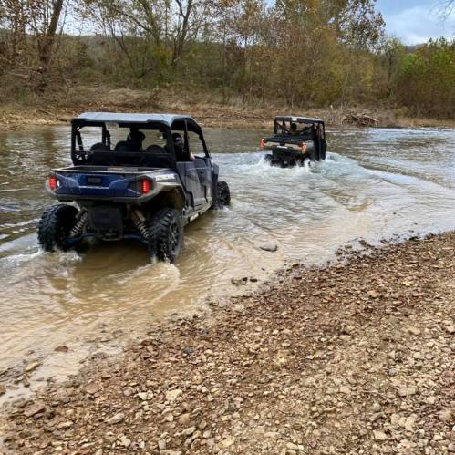 Two off-road vehicles driving through a shallow river, surrounded by trees and rocky terrain.