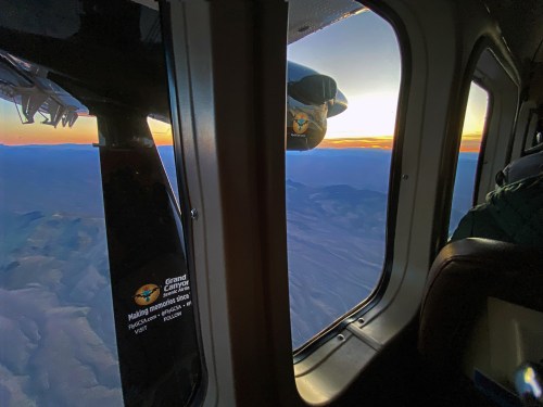 View from a helicopter window showing a sunset over mountains and a propeller in the foreground.