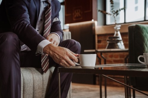A man in a suit sits on a couch, holding a coffee cup near a glass table in a stylish, modern setting.
