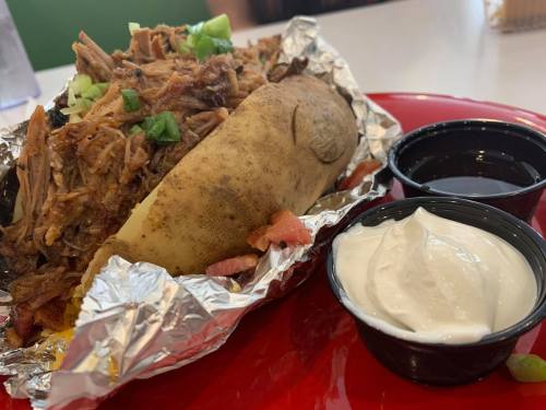 A loaded baked potato topped with pulled pork, green onions, and served with sour cream and barbecue sauce on the side.