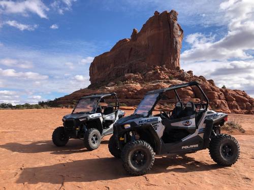 Two off-road vehicles parked on red rock terrain with a large rock formation and blue sky in the background.