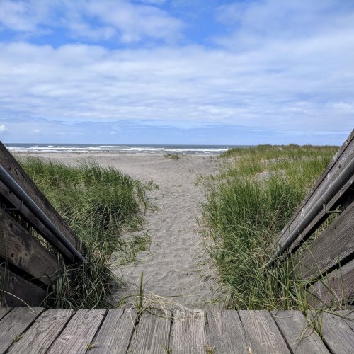 A wooden pathway leads to a sandy beach with grass on either side, under a partly cloudy sky.