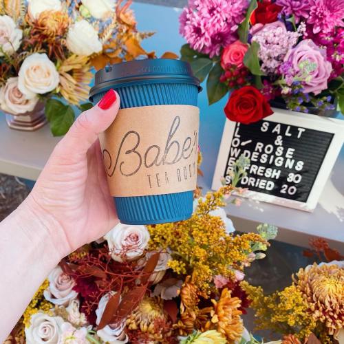 A hand holds a coffee cup with "Babe's Tea Room" label, surrounded by colorful flowers and a decorative sign.