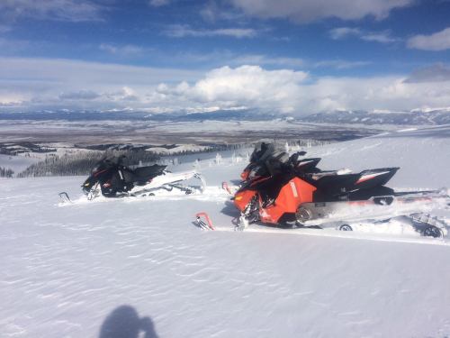 Two snowmobiles parked on a snowy mountain slope, with a scenic view of valleys and distant mountains under a blue sky.