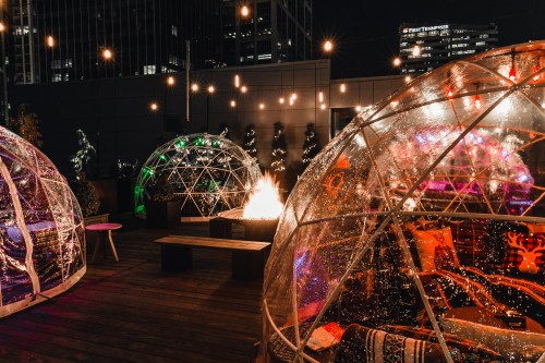 Cozy outdoor igloos with warm lighting around a fire pit, set against a city skyline at night.