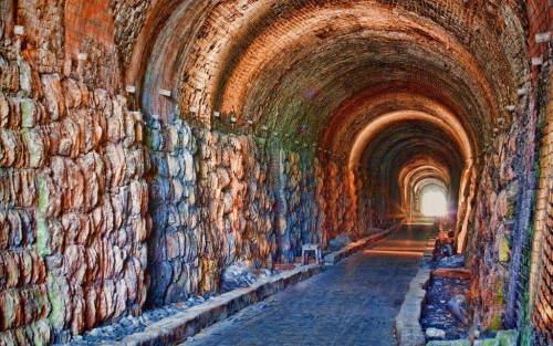 A long, arched tunnel with stone walls, leading to a bright light at the far end. The ground is uneven and rocky.