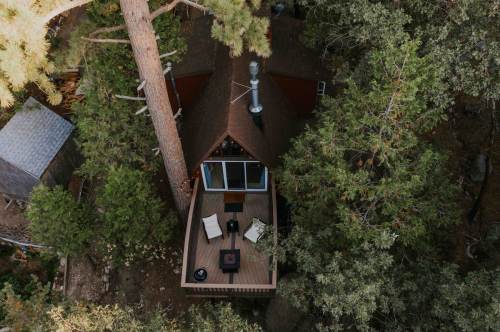 Aerial view of a cozy cabin surrounded by tall trees, featuring a deck with seating and a fire pit.