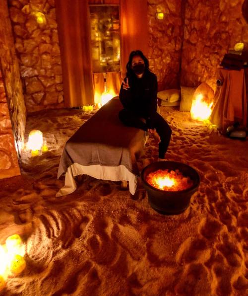A person in a mask sits on a bed in a dimly lit room with glowing salt lamps and a fire pit.